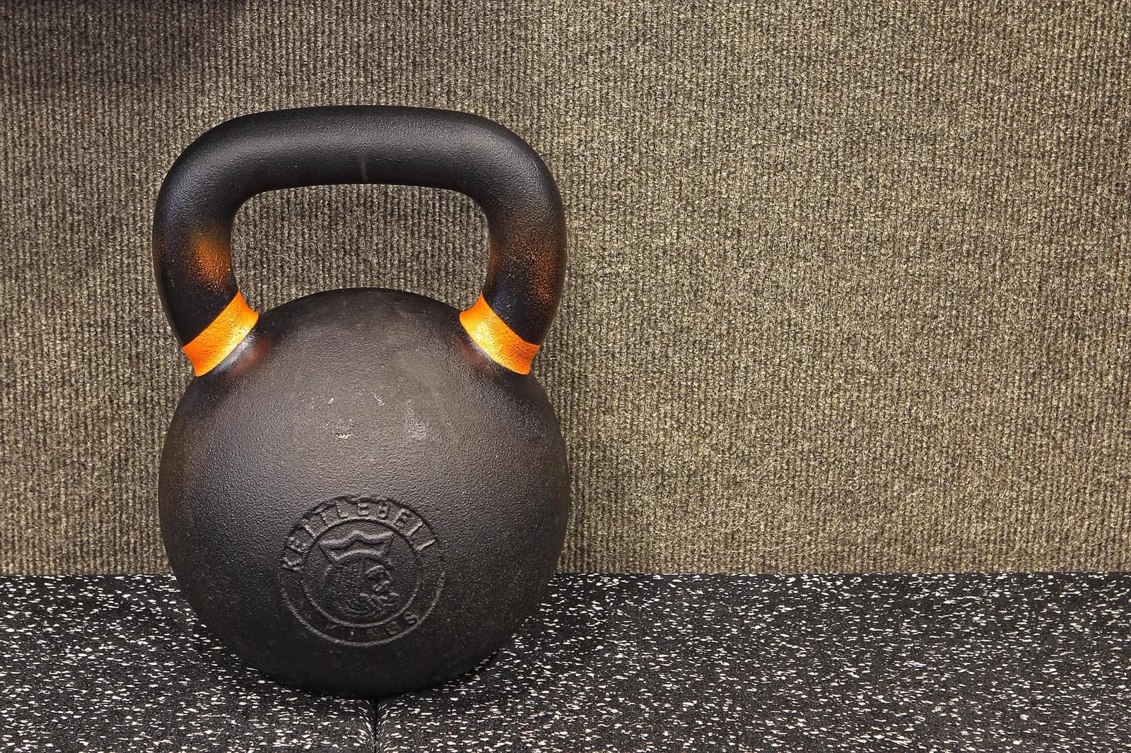 A cast-iron kettlebell with orange banding on its handle, standing on speckled rubber flooring against a carpeted wall.