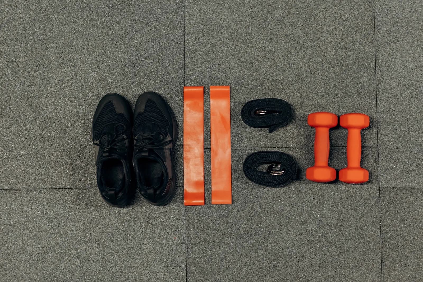 Black trainers, two orange resistance bands, a pair of lifting straps and two orange dumbbells laid out in a row on grey rubber gym tiles.