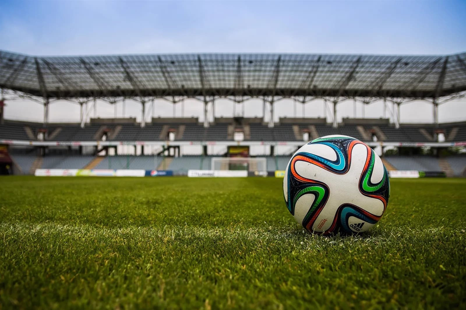 A match ball resting on the grass of an empty stadium, with covered stands rising behind it.