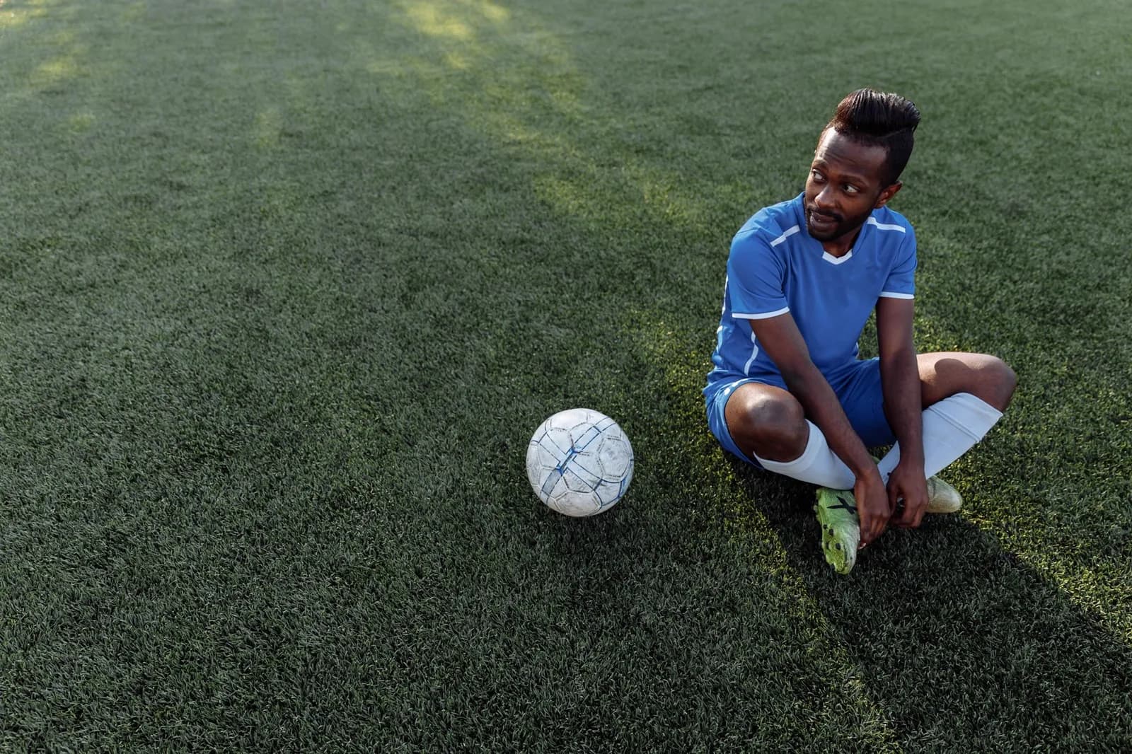 A footballer in a blue kit sitting cross-legged on sunlit artificial turf beside a ball, reaching down to a boot.