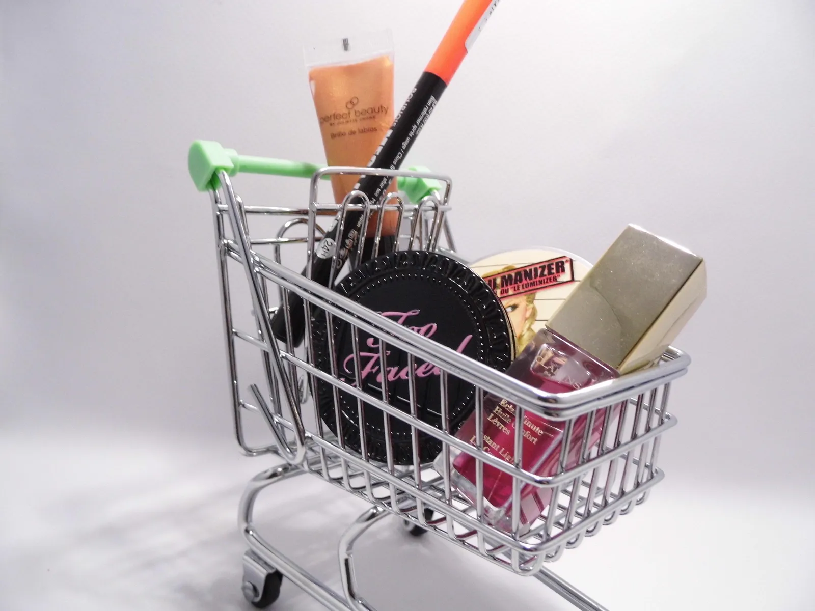 A miniature chrome shopping trolley filled with cosmetics — a lip gloss, an eyeliner pencil, a compact and a nail varnish.
