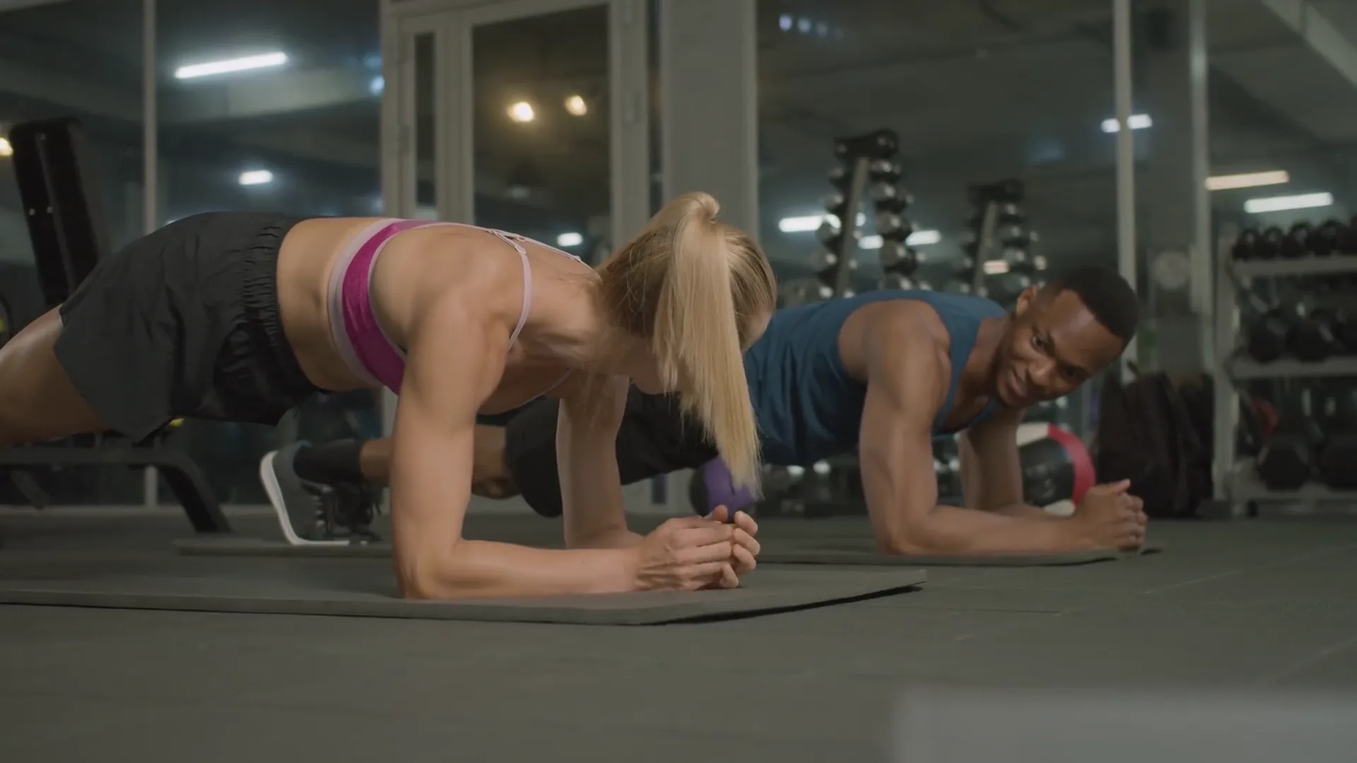 Two athletes holding a forearm plank on mats in the TAY gym, weight racks behind them.